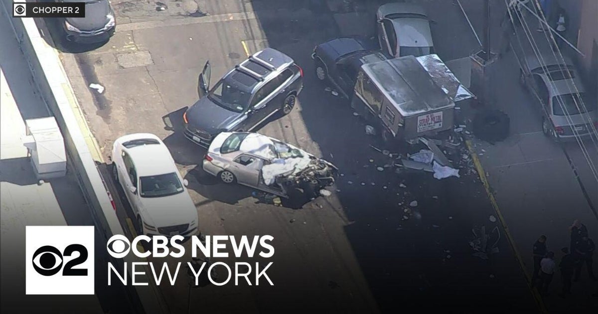 Aerial photo showing several damaged vehicles and debris on a city street in Queens. A silver sedan with its front end crushed sits near a box truck and other cars; doors are open and people, including emergency responders, are visible on the sidewalk.