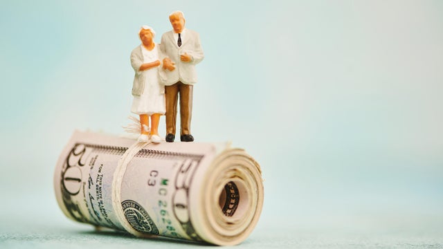 Miniature figures of elderly couple standing on top of a wad of fifty dollar bills 