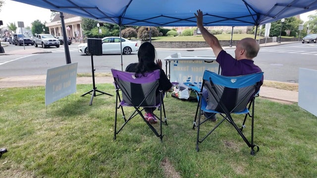 Will Thilly sits under a tent and waves at a passing car 