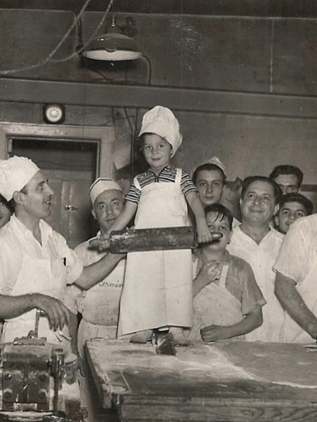 A young Vincent Termini wearing an apron and bakers' hat holds a rolling pin, surrounded by other bakers