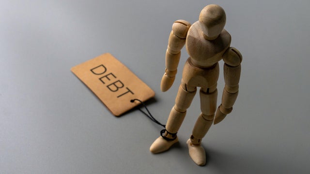A wooden figure stands with one ankle tied by a string to a tag labeled DEBT. 