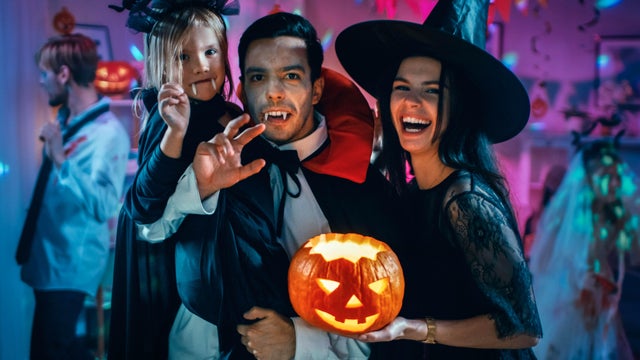 Halloween Costume Party: Father Count Dracula Holds Little Bat Girl Daughter and Hugs Witch Wife for a Happy Family Portrait. In the Background Monsters Having Fun 