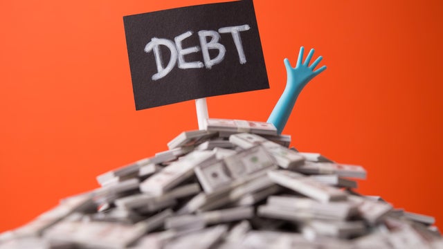 Pop out hand from heap US dollar banknotes and DEBT label white wall background. 