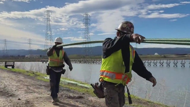 Two people in safety vests carry rebar to a pond restoration project site 