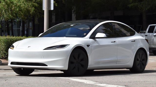 A portrait of a white Tesla Model 3 EV cruising in a downtown district 