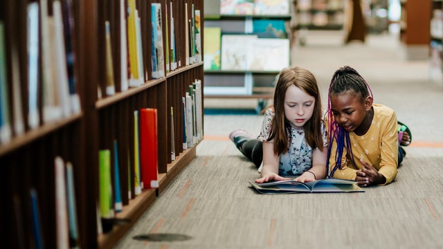 Two young girls reading a book on the floor of library 
