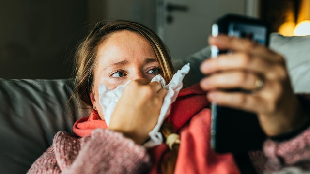 Ill woman lying on the sofa and coughing and sneezing while using mobile phone for digital sick note. 