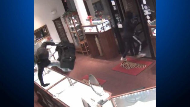 A person in black clothing jumping over a jewelry store counter, carrying a black bag 