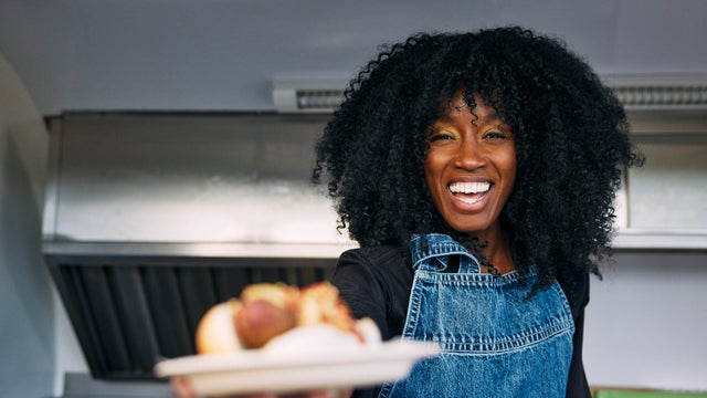Portrait of happy mature female owner with curly hair holding plate of vegetarian hot dog at food truck 
