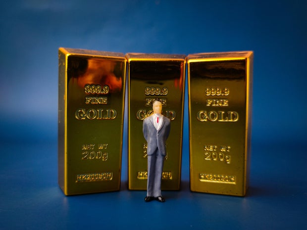 This image depicts a miniature figure of a businessman standing in front of three gold bars, symbolizing business and financial concepts 