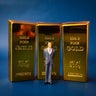 This image depicts a miniature figure of a businessman standing in front of three gold bars, symbolizing business and financial concepts 