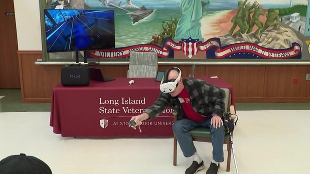 Man with cane sits in chair and uses VR goggles to bowl 