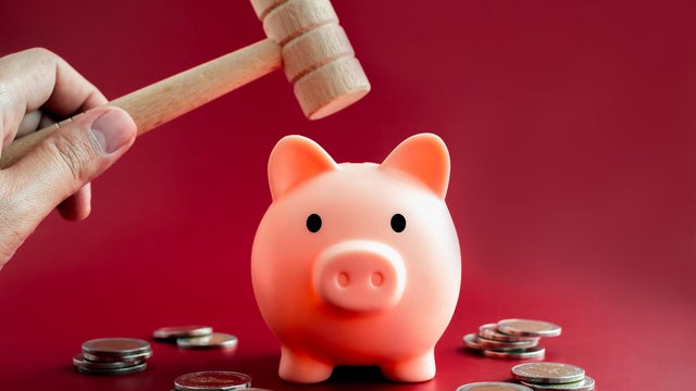 The pink piggy bank, front view is about to be smashed with a wooden hammer in hand over the coins piles on red background. 