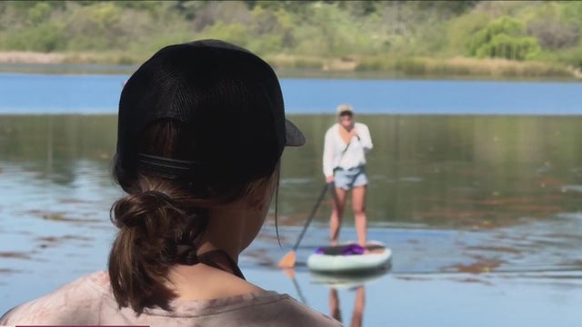 A woman looks out at a lake as someone on a paddleboard approaches 