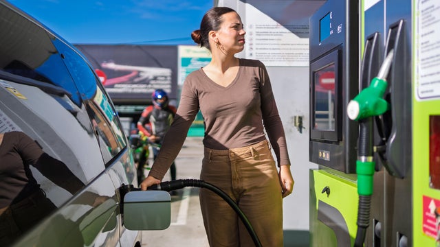 Woman contemplating rising gas prices and environmental impact while refueling car on sunny day. The intersection of transportation, energy, and the environment in everyday life 