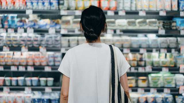 Rear view of young Asian woman doing grocery shopping, standing in front of a shelf full of fresh dairy products in the dairy aisle and thinking which product to choose from in a supermarket. Concept of customer awareness 