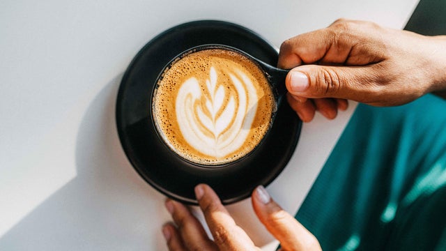 A person hands holds a cappuccino cup with elegant latte art on a black saucer, set against a bright white table. 