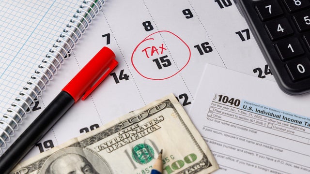 Close up of a calendar with a red circle on the 15th and color Form 1040 next to a hundred dollar bill and modern office supplies shot. 