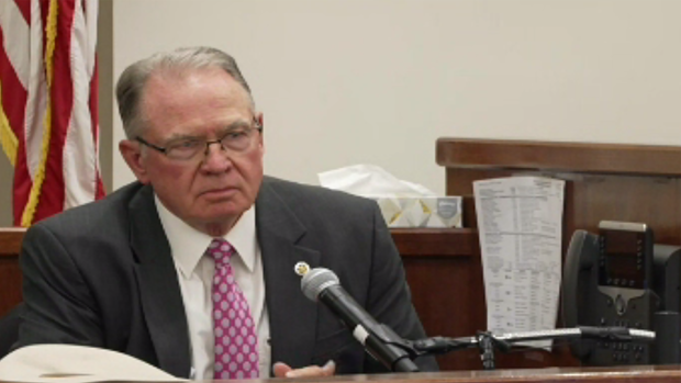 Former sheriff Lane Akin sits in a brown courtroom witness stand. He's wearing a black sport coat, white button-up shirt, and pink patterned tie. 