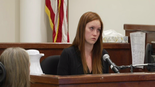 Ashley Strand at brown courtroom witness stand. She's wearing a black blouse and has red hair, freckles, and blue eyes. 