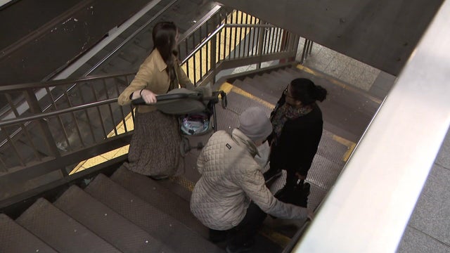 Woman with a stroller walks down subway stairs while another woman with heavy bag walks up 