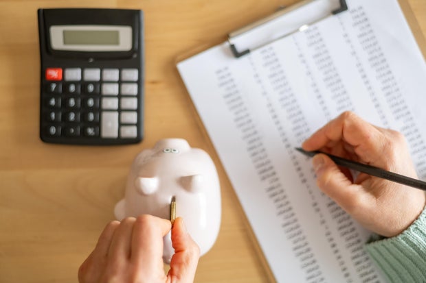 Accountant inserting a coin into piggy bank and calculating expenses with calculator and spreadsheet on a wooden desk, managing home budget and savings 