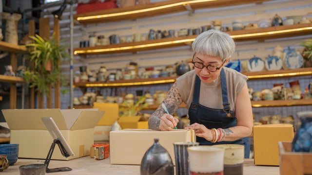 Senior artist women, owner of  pottery shop packing product in boxes, writing shipping address on box for delivery. 