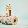 Miniature figures of elderly couple standing on top of a wad of fifty dollar bills 