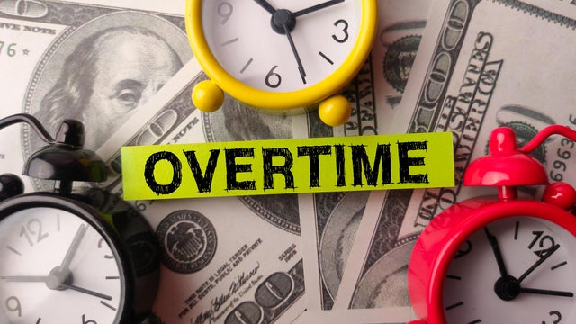 Alarm clock and sticky note with the word OVERTIME on a one hundred dollars 