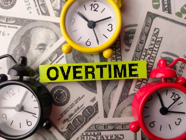 Alarm clock and sticky note with the word OVERTIME on a one hundred dollars 