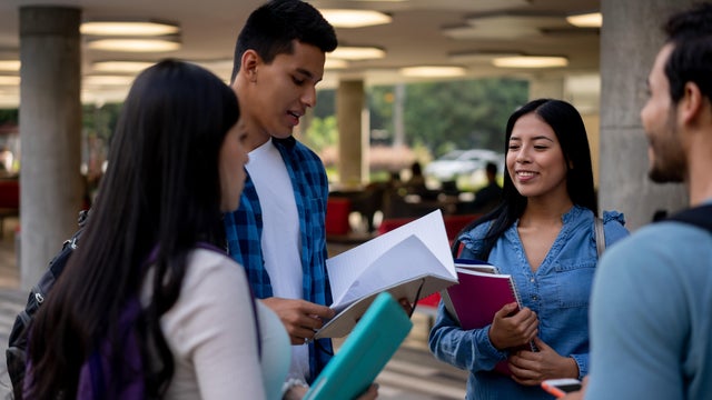Group of college buddies standing next to partner reading something on notebook at college 