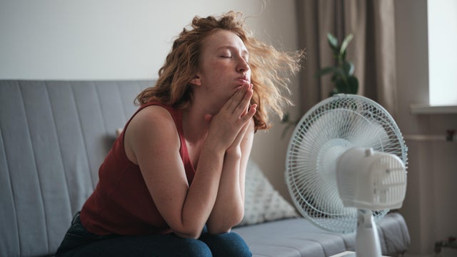Suffering from summer heat at home, exhausted young woman sitting near electric fan enjoying fresh air, cooling down during heatwave 