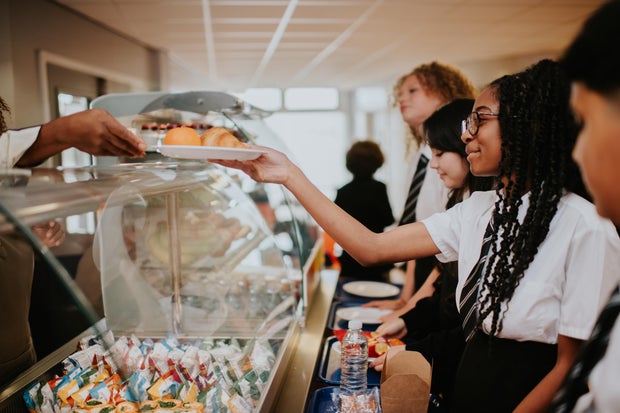 A group of high school students wait in line in a school cafeteria at lunch time. They hold trays whilst they make their food choices. A dinner lady passes a girl a plate of food.