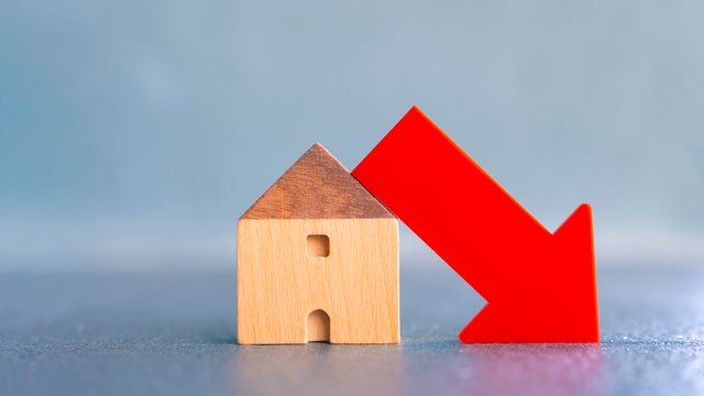 Wooden house model with red down arrow showing decrease in real estate market, home value, or housing demand. Concept of economic recession, property price drop, or mortgage crisis. 