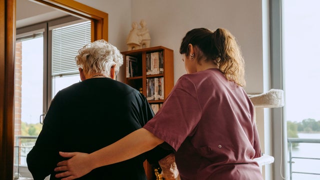 Rear view of female caregiver supporting senior woman while walking in apartment 