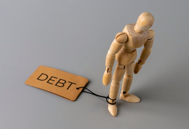 A wooden figure stands with one ankle tied by a string to a tag labeled DEBT.