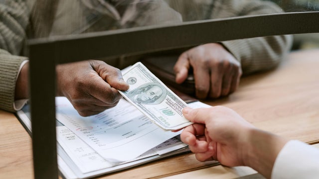 Middle Aged Black Man Handing Cash to Caucasian Bank Teller at Branch Counter 